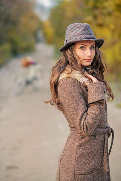 Beautiful girl in autumn coat and hat standing on the dirt road with ladies bicycle and autumn trees on the background.