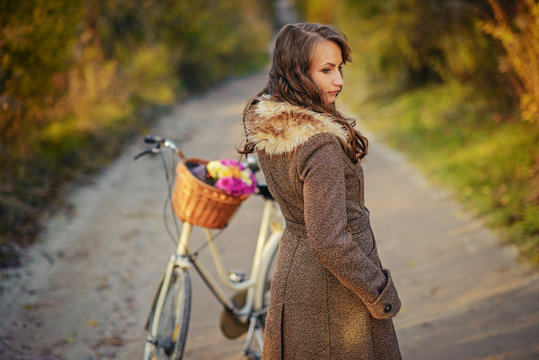 Beautiful girl in autumn coat standing near the ladies bicycle with pink and yellow flowers in a basket with autumn trees behind.