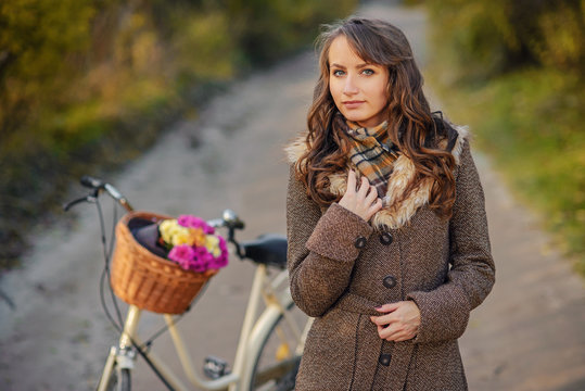 Beautiful girl in autumn coat standing near the ladies bicycle with pink and yellow flowers in a basket.