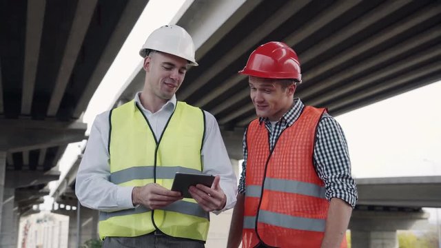 4K shot of Two young workers in red and yellow vests and hard hats lookig at tablet computer and discusses a project while standing under the bridge construction. Movement stabilized