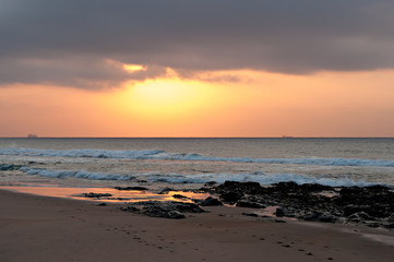 The sun rising into a bank of cloud, with rocks and beach in the foreground and ships on the horizon.