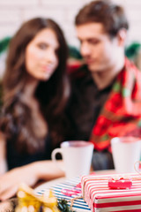 Christmas present, happy couple blurred background. Focus on red new year gift, family with hot chocolate cups backdrop. Holiday, party, winter, coziness concept