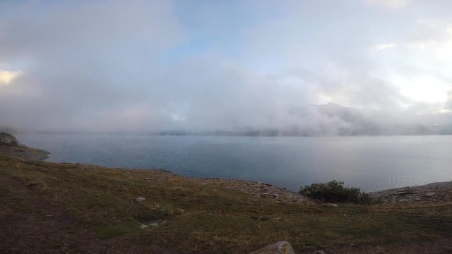 Time lapse Mont Cenis, France, artificial lake at the mountain pass. Fog is covering the lake during autumn season. First fresh snow covers the mountain tops and the sun rays pass through clouds