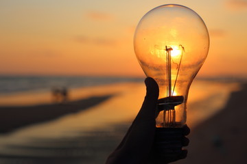 hand holding a electric light bulb and sun at beach sunset sky