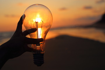 hand holding a electric light bulb and sun at beach sunset sky