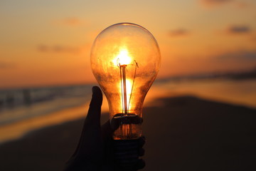 hand holding a electric light bulb and sun at beach sunset sky