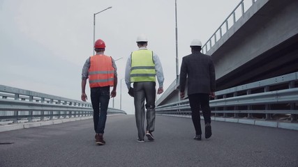 4K shot of Diverse group of three male construction engineers in white hard hats with reflective jackets walking on highway ramp. Rear view on road along bridge.
