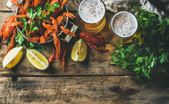 Two mugs of wheat beer and boiled crayfish in pan served with with lemon and parsley over rustic wooden background, top view, copy space, horizontal composition