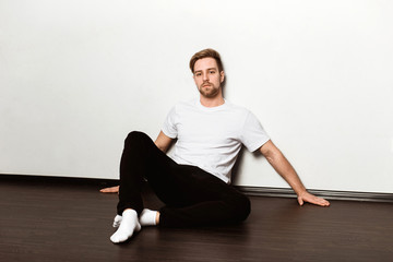 A young caucasian blond man in a white t-shirt and black trousers sitting on the floor