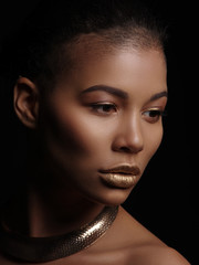 Fototapeta premium Portrait of an extraordinary beautiful naked african american model with perfect smooth glowing skin, make up, full golden lips, shaved haircut and gold jewelry on her neck, dark background