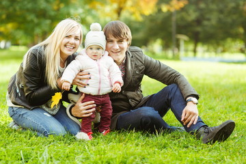 Naklejka na ściany i meble Cheerful parents with daughter sitting on grass in autumn park