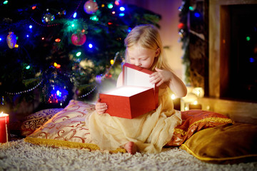 Happy girl opening Christmas gifts by a fireplace