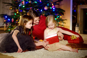 Fototapeta na wymiar Young mother and her two daughters unwrapping Christmas gifts by a fireplace