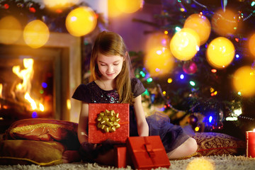 Happy girl opening Christmas gifts by a fireplace