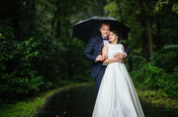 Fototapeta premium Laughing bride clung to the groom in the rain with an umbrella, standing on the asphalt track in the rain