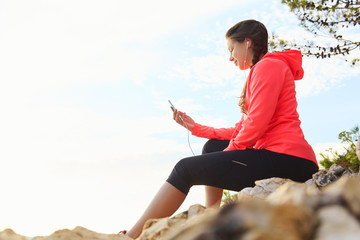 Active young woman listening to music. Runner resting from routine exercise. Happy jogger relaxing at the beach after run and sitting on the stones.