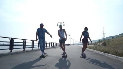 Silhouette three friends skateboarders on the waterfront. Lower part of the body two young gues and one girl on longboard and ska