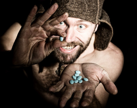 Dark Portrait Of Scary Evil Sinister Bearded Man With Smirk, Holds In Palm Of A Bunch Of Pills. Strange Russian Man With A Naked Torso And A Woolen Hat