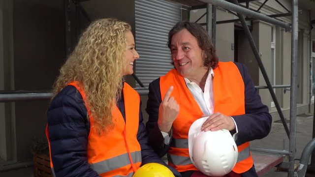 Male And Female Construction Workers Sitting On Scaffolding And Laughing Together