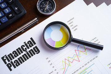 "Financial Planning" text on paper sheet with magnifying glass on chart, dice, spectacles, pen, laptop and blue and yellow push pin on wooden table - business, banking, finance and investment concept