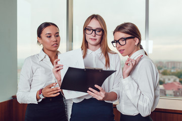 One Asian and two European business women standing against the window  study the documents