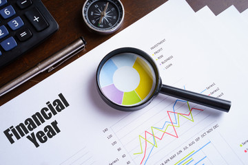 "Financial Year" text on paper sheet with magnifying glass on chart, dice, spectacles, pen, laptop and blue and yellow push pin on wooden table - business, banking, finance and investment concept
