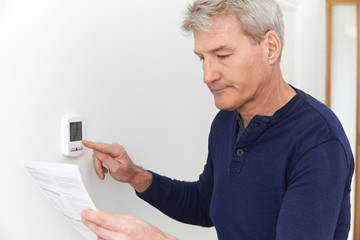 Worried Mature Man With Bill Turning Down Heating Thermostat