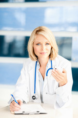 Attractive blonde female woman doctor nurse practitioner in lab coat and stethoscope with mobile phone in hospital clinic medical center office