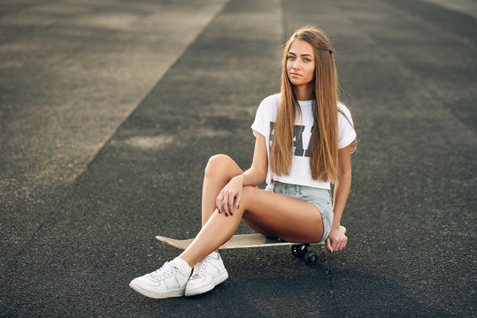 Beautiful young and sexy girl with long hair in a white T-shirt, short shorts and sneakers sitting on a skateboard