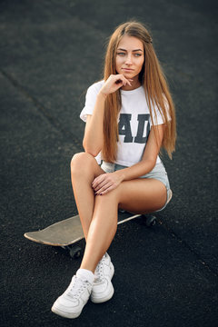 Cute beautiful young and sexy girl in a white T-shirt, short shorts and sneakers sitting on a skateboard