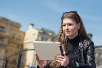 Young woman tourist is reviewing city map via app in touch pad, while is waiting transport on a bus stop. Beautiful female student is searching information in internet via portable digital tablet