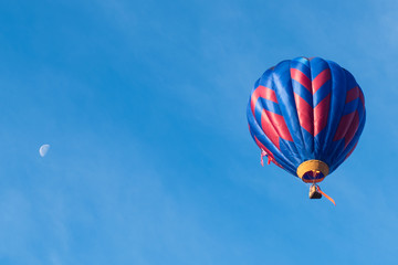 This is a photo of a beautiful hot air balloon slowly sailing through a calm blue sky with the moon in the background.