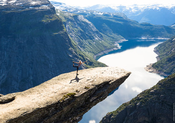 Young woman doing yoga on Trolltunga. Happy girl enjoy beautiful lake and good weather in Norway.