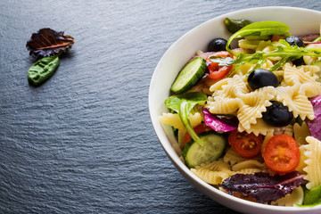 Pasta salad made from farfalle, red onion, cherry tomatoes, fresh cucumbers, black olives, mix of green salad leaves. White bowl on black stone background.