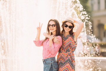 Two happy girls stand by fountain and possing on summer streets