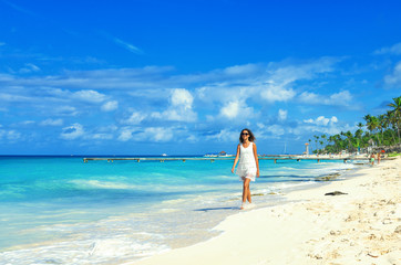 Fototapeta na wymiar Young woman walking along white sand tropical beach. Beautiful girl in white dress on the beach. Travel and vacation. Freedom concept.