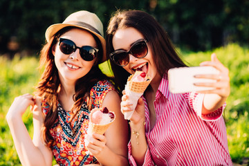 Portrait of two young women standing together eating ice cream and taking selfie photo on camera in summer street.