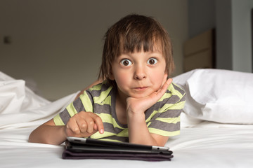 Little boy with digital tablet rests, surprised on the bed on home interior background