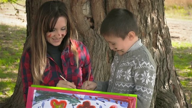 Happy son and mother enjoy beautiful pictures and kiss. Mother and son paint on the paper. Young mother with her son drawing on the easel under a big tree