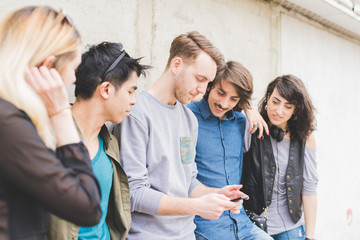 Knee figure of a group of young multiethnic friends leaning on a wall chatting to each other having fun using smartphone - social network,technology, communication, technology concept