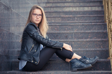 Teen girl with long blond hair sitting on the marble steps. Beautiful young girl in glasses and black leather jacket.