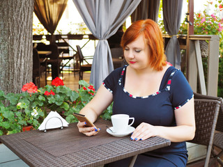 Portrait of attractive plus size redhead woman sitting in a cafe in city park with cup of coffee and checks her smart phone