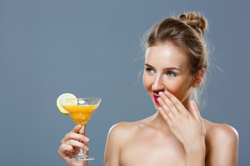 Beautiful blonde naked girl smiling, holding cocktail over grey background.