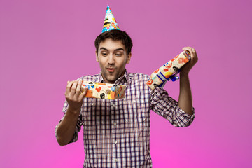 Happy young handsome man opening birthday gift over purple background.