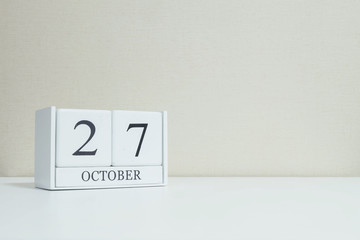 Closeup white wooden calendar with black 27 october word on blurred white wood desk and cream color wallpaper in room textured background with copy space , selective focus at the calendar