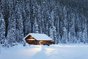 A snow covered log cabin on a snow covered lakeshore surrounded by evergreen trees at dusk;Lake louise alberta canada