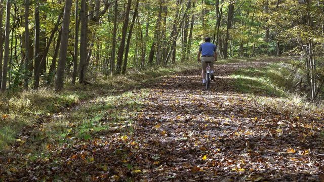 Senior caucasian adult man cycling away from the camera on a leaf covered forest trail near Morgantown illustrating the importance of exercise to older people