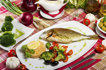 Fried gilt-head bream on a plate with a side dish of broccoli, cherry tomatoes, rice, olives and lemon served beside fresh vegetables.
