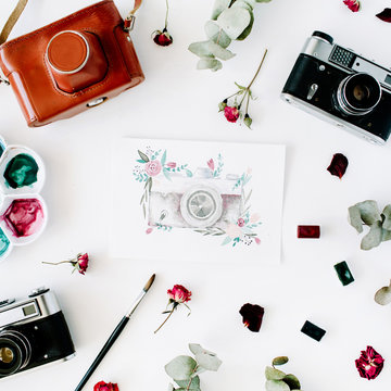 Flat lay. Artist workspace with vintage retro photo camera, and watercolor painted camera, red roses and eucalyptus. Top view arrangement