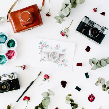 Flat lay. Artist workspace with vintage retro photo camera, and watercolor painted camera, red roses and eucalyptus. Top view arrangement